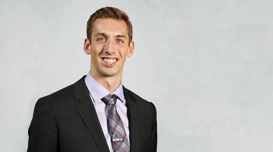 Rick Gevis, BBA '18, MSA '19 smiles in a suit against a gray backdrop.